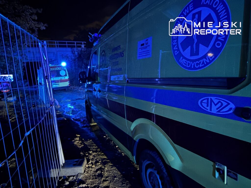 Karetka pogotowia stoi nocą za metalowym ogrodzeniem, oświetlona niebieskim światłem.