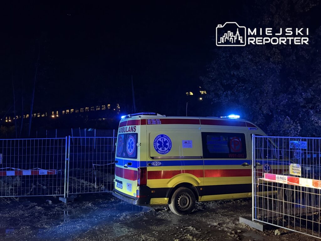 Karetka pogotowia z włączonymi niebieskimi światłami stoi nocą za metalowymi barierkami przy torach kolejowych.