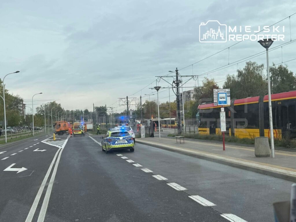Radiowóz i samochód ratowniczy na ulicy przy przystanku tramwajowym, obok stoją tramwaje i służby ratunkowe.