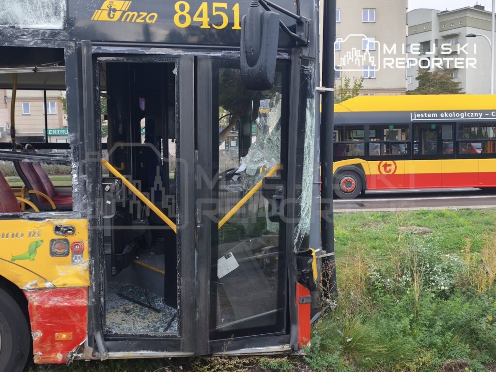Uszkodzony autobus z rozbitą przednią szybą i wybitymi drzwiami stoi przy trawniku; w tle drugi autobus.