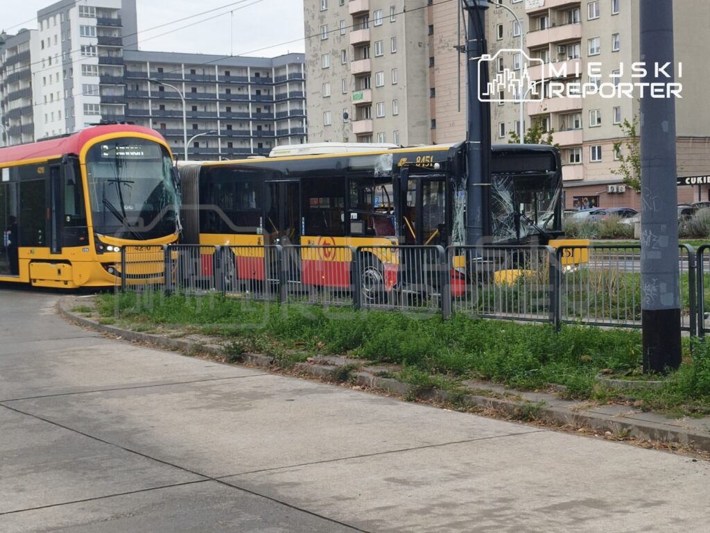Żółto-czerwony autobus stoi obok tramwaju za metalowym ogrodzeniem, na tle bloków mieszkalnych.