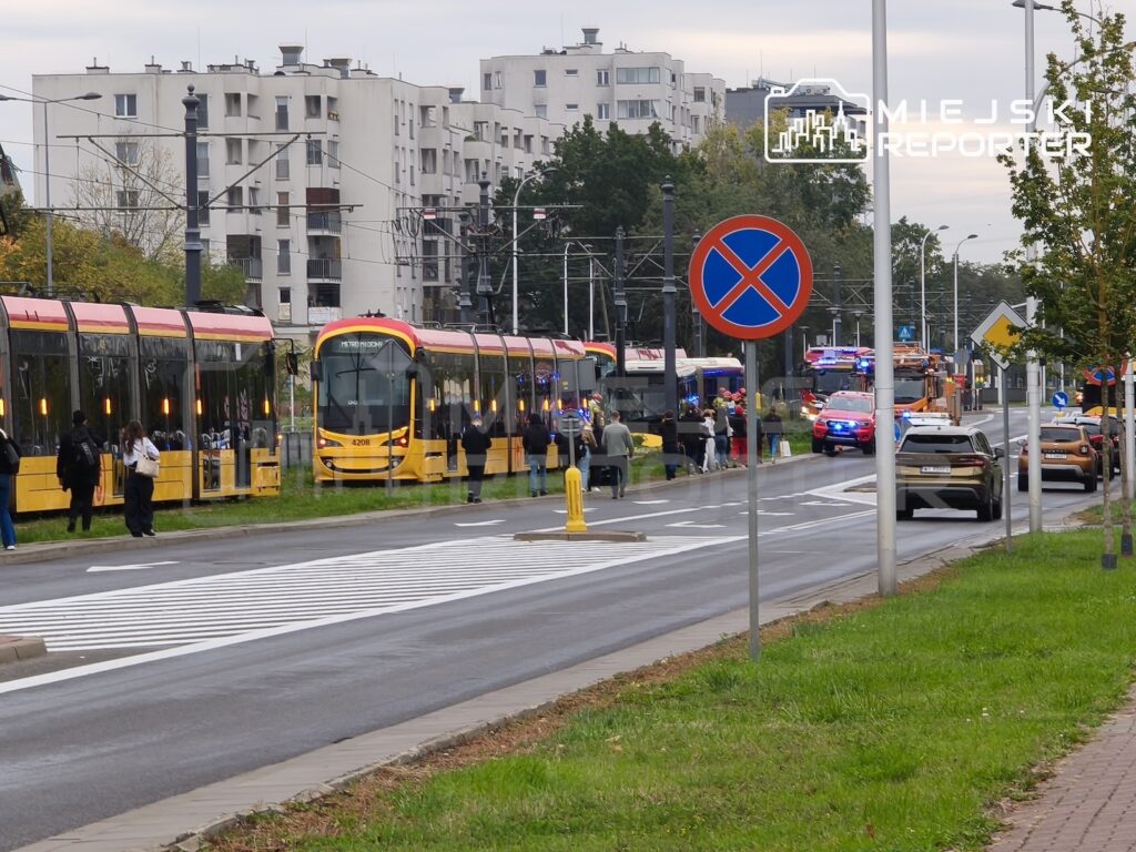 Żółte tramwaje stoją przy torowisku, a na ulicy obok widać radiowozy z włączonymi światłami i zatrzymane auta.