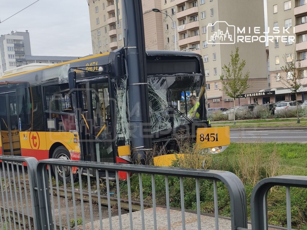Uszkodzony żółto-czerwony autobus z rozbitą przednią szybą stoi przy torowisku, obok słupa i bloków mieszkalnych.
