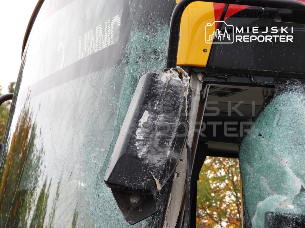 Zbliżenie na uszkodzony autobus z rozbitą przednią szybą i pękniętym lusterkiem bocznym, widok z zewnątrz.