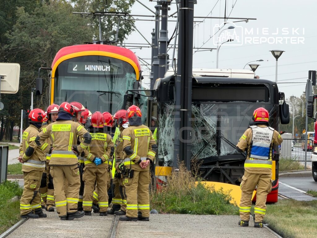 Strażacy w odzieży ochronnej oglądają uszkodzony autobus stojący przed tramwajem na torowisku.