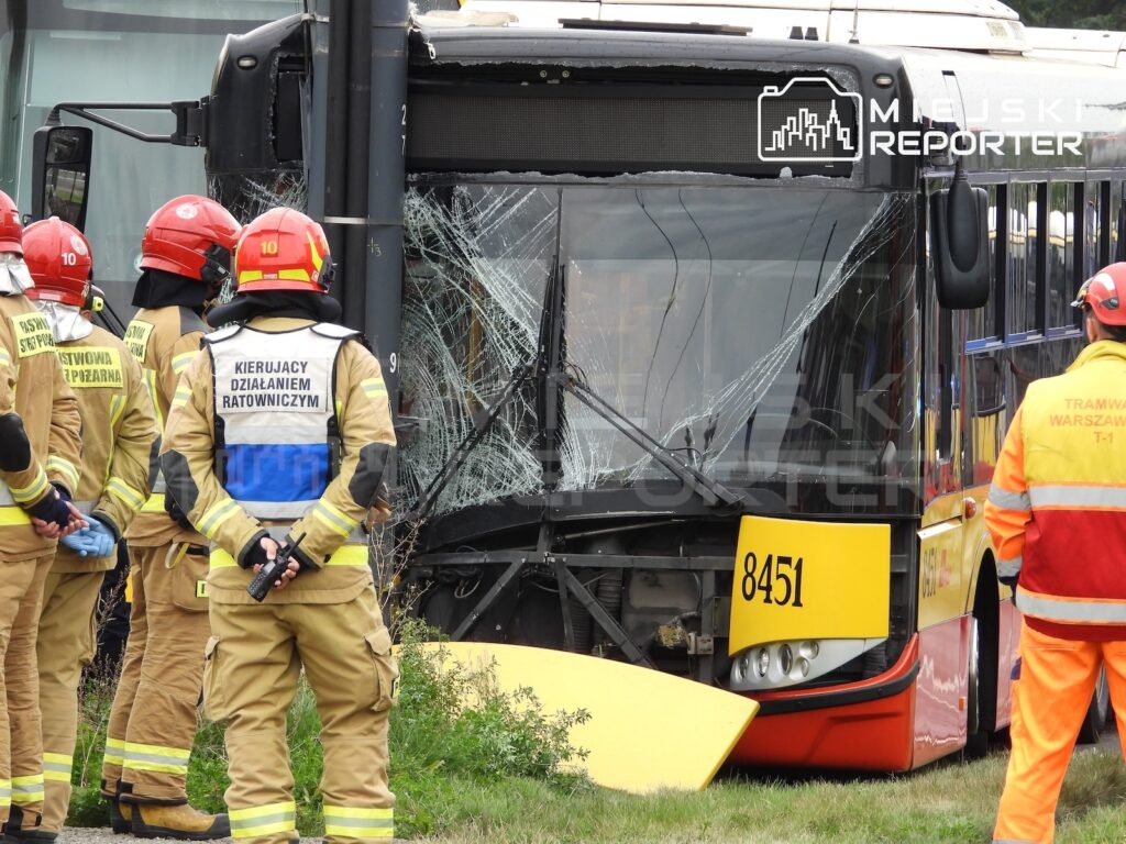 Strażacy oglądają autobus z rozbitą przednią szybą; obok stoi pracownik w pomarańczowym stroju.