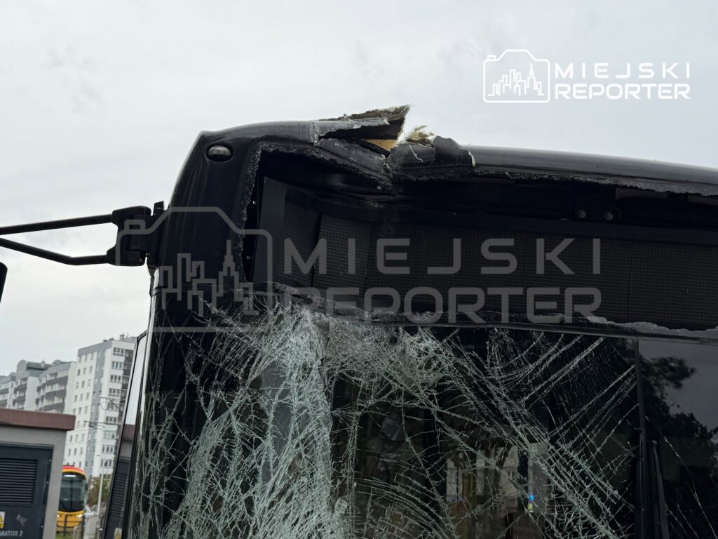 Uszkodzony przód autobusu z rozbitą szybą, widocznym pęknięciem karoserii i oderwanym fragmentem dachu na tle bloków.