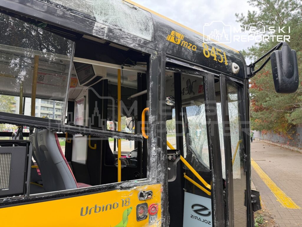 Żółty autobus z rozbitą szybą i uszkodzonymi drzwiami stoi przy przystanku tramwajowym.