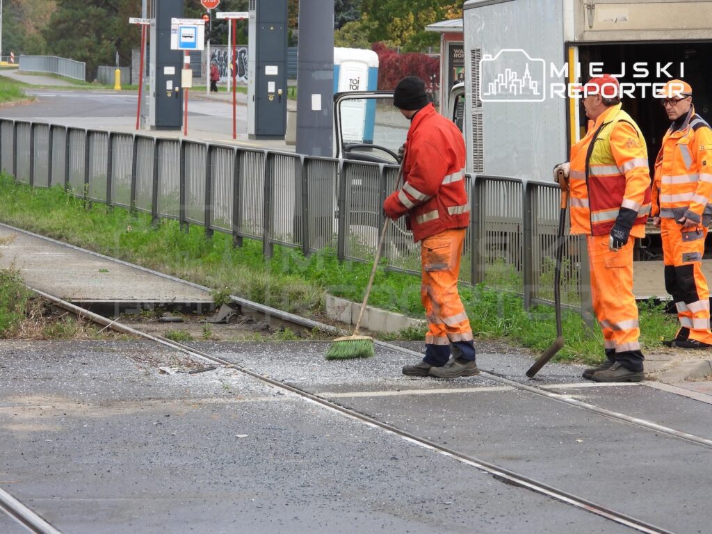 Trzech pracowników w pomarańczowych strojach porządkowych sprząta przy torowisku tramwajowym i przejściu przez tory.