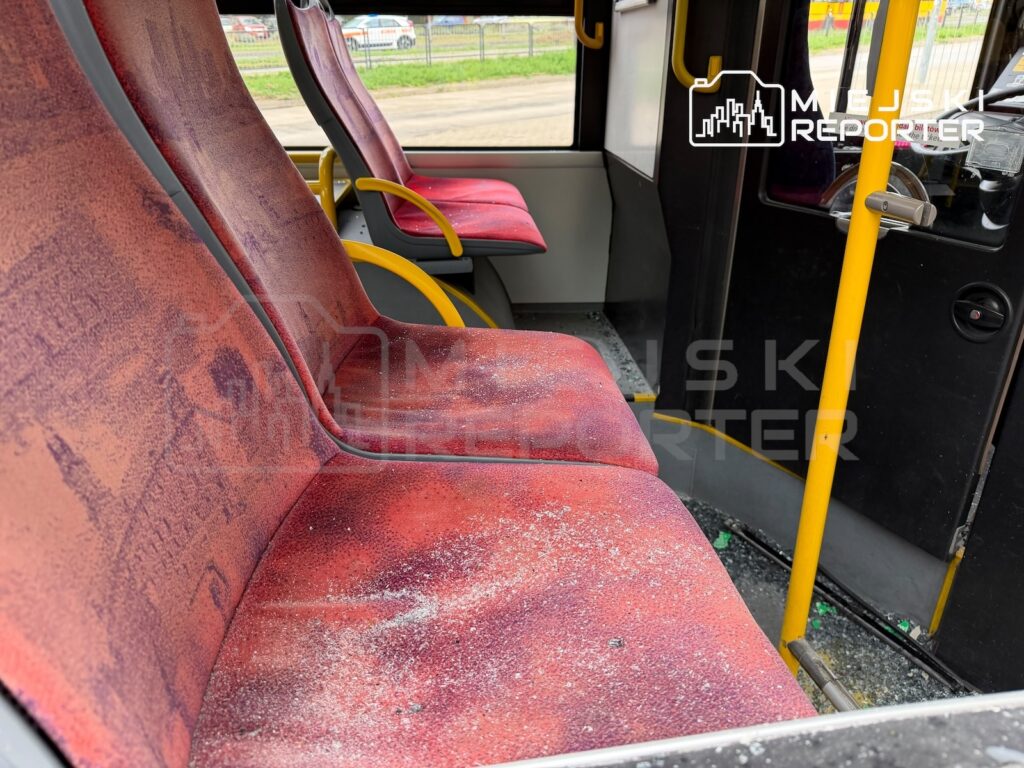 Zabrudzone czerwone siedzenia i rozbite szkło na podłodze we wnętrzu autobusu miejskiego.