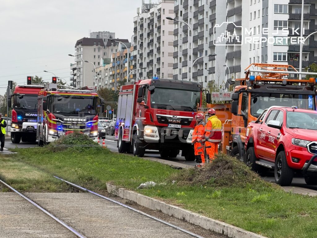 Strażacy i służby techniczne pracują przy torowisku, obok wozów strażackich i samochodów służbowych na miejskiej ulicy.