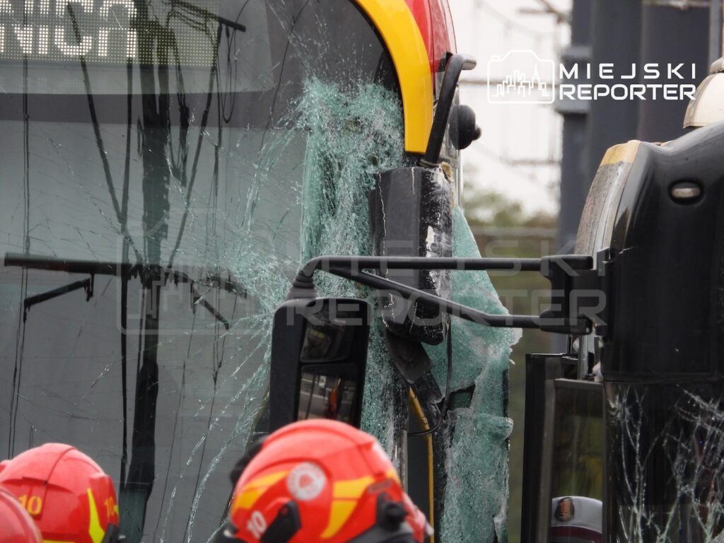 Przednia szyba żółto-czerwonego autobusu jest rozbita, a obok stoją strażacy w czerwonych hełmach.