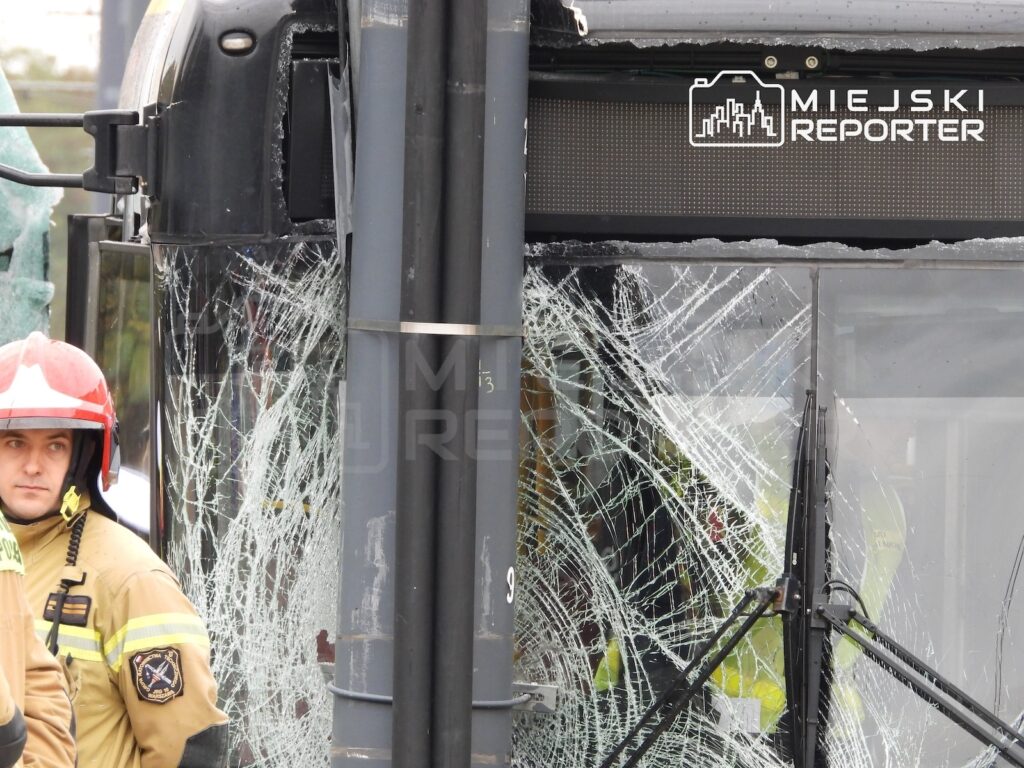 Uszkodzony autobus z rozbitą przednią szybą, obok strażak w hełmie ochronnym.