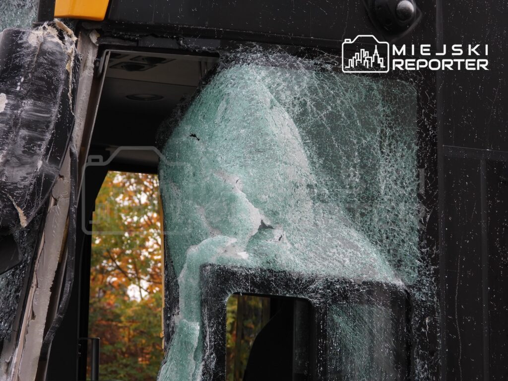Popękana szyba w drzwiach autobusu, z widocznymi odłamkami szkła i uszkodzonym obramowaniem.