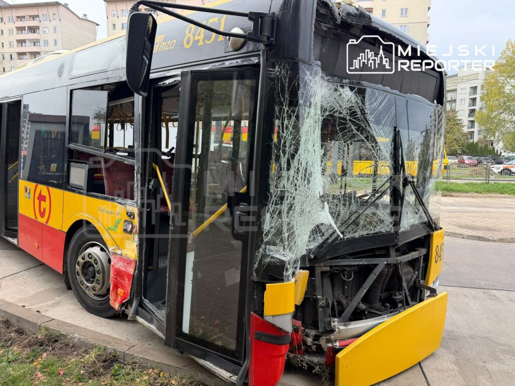 Uszkodzony autobus miejski z rozbitą przednią szybą i zniszczonym przodem stoi na jezdni przy zabudowie mieszkalnej.