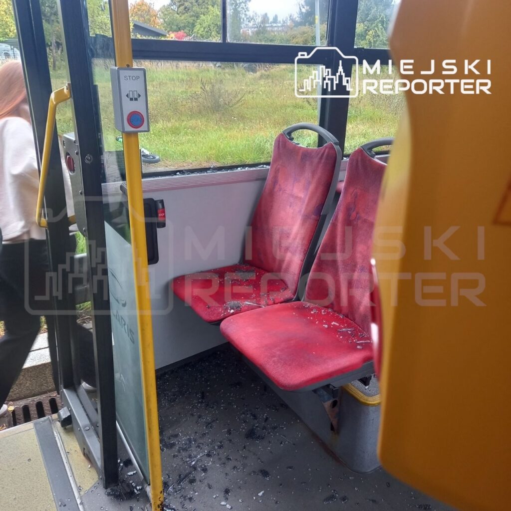Rozbita szyba i rozsypane odłamki szkła przy czerwonych siedzeniach w autobusie miejskim.