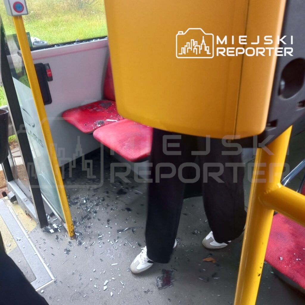 Rozbite szkło leży na podłodze autobusu przy wejściu i czerwonych siedzeniach.