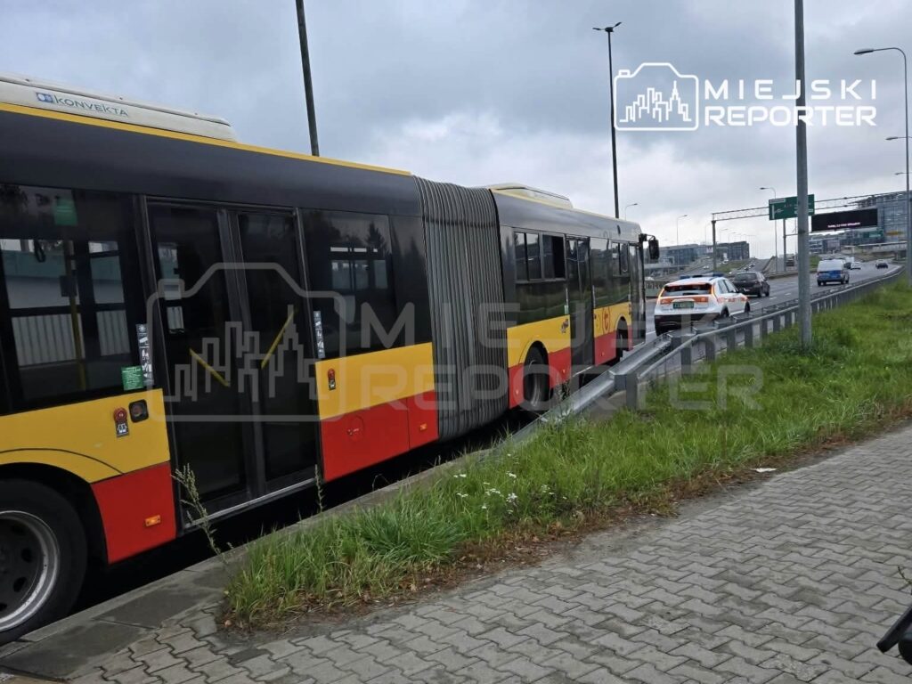 Przegubowy autobus miejski w żółto-czerwonych barwach stoi przy drodze, za nim znajduje się samochód służb.