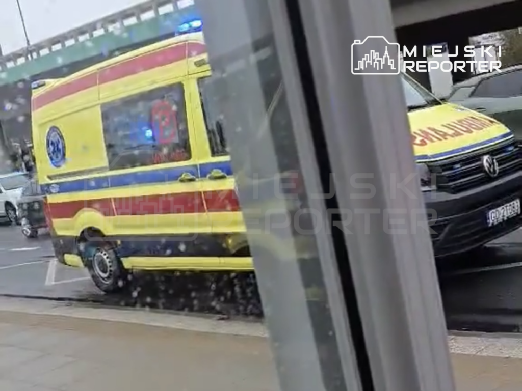 Żółty ambulans zaparkowany przy chodniku, częściowo zasłonięty przez słup.