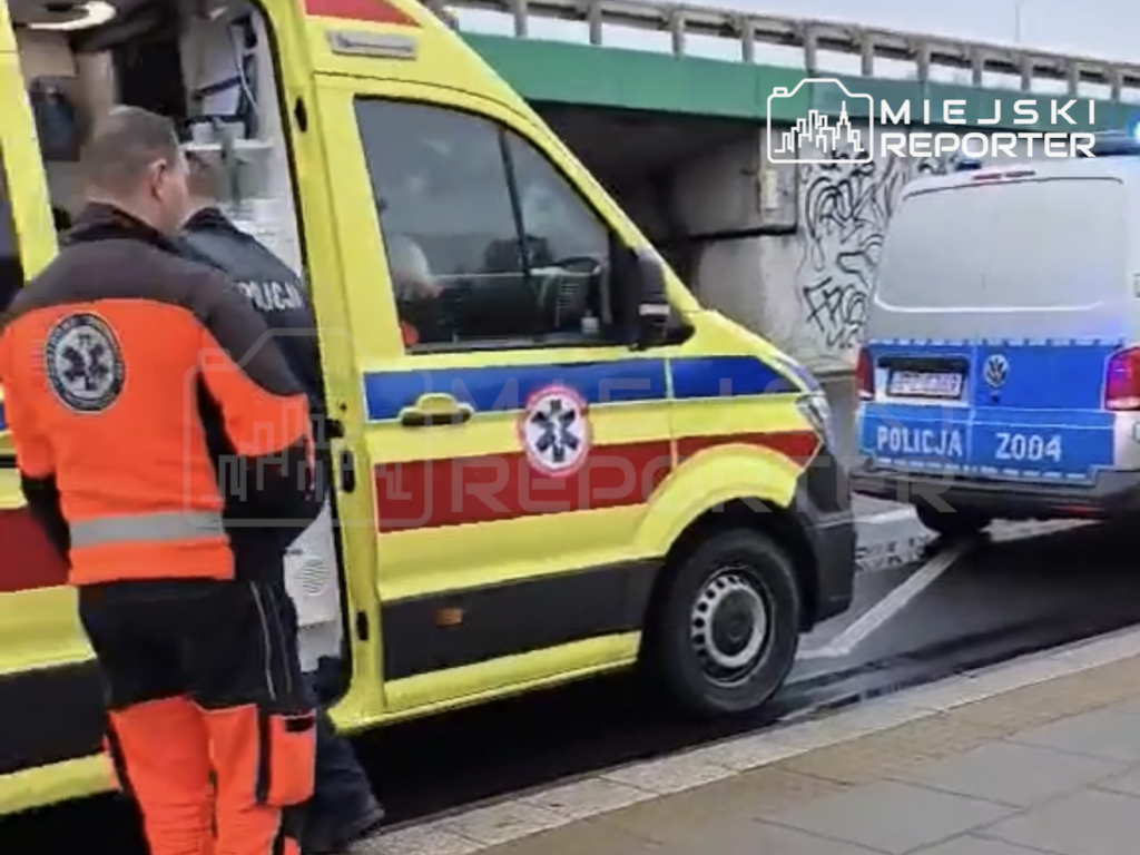 Ratownik medyczny stoi przy otwartych drzwiach żółtej karetki, obok zaparkowanego radiowozu policji.