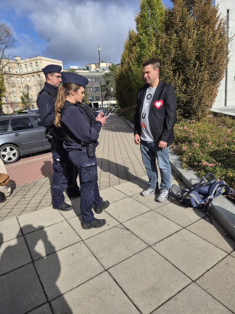 Policjantka sprawdza telefon, stojąc obok policjanta i mężczyzny na chodniku przy zaparkowanym samochodzie.