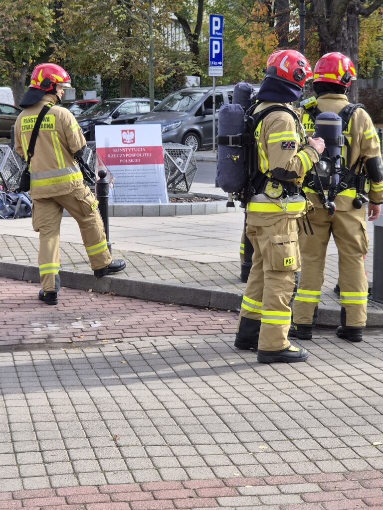 Strażacy w hełmach i ubraniach ochronnych stoją przy makiecie z napisem „Konstytucja Rzeczypospolitej Polskiej” na chodniku.