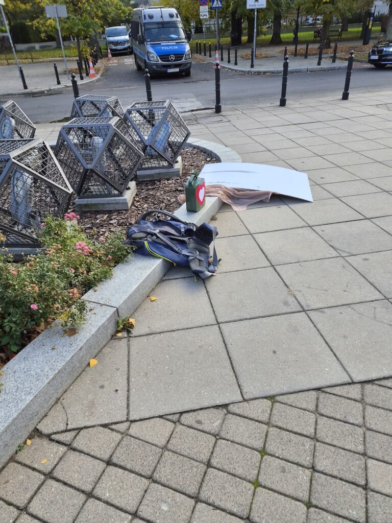 Na chodniku leżą przewrócona biała plansza, plecak i zielony pojemnik; w tle stoi policyjny bus.