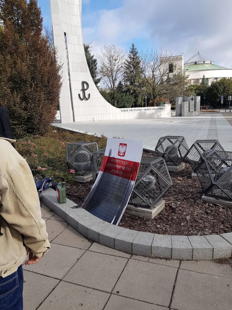 Makieta Konstytucji RP oparta o kamienny mur przy pomniku Armii Krajowej; obok stoją metalowe konstrukcje i osoba w jasnej