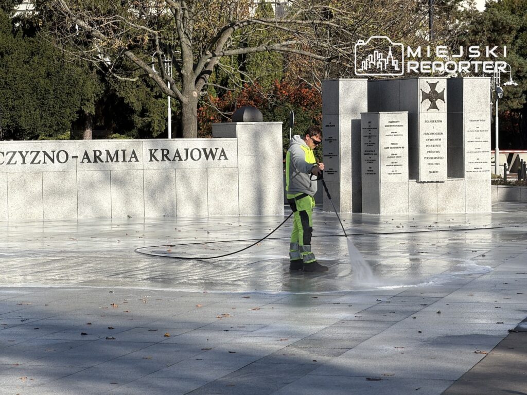 Pracownik w odblaskowym ubraniu myje wodą kamienny plac przed pomnikiem z tablicami pamiątkowymi.