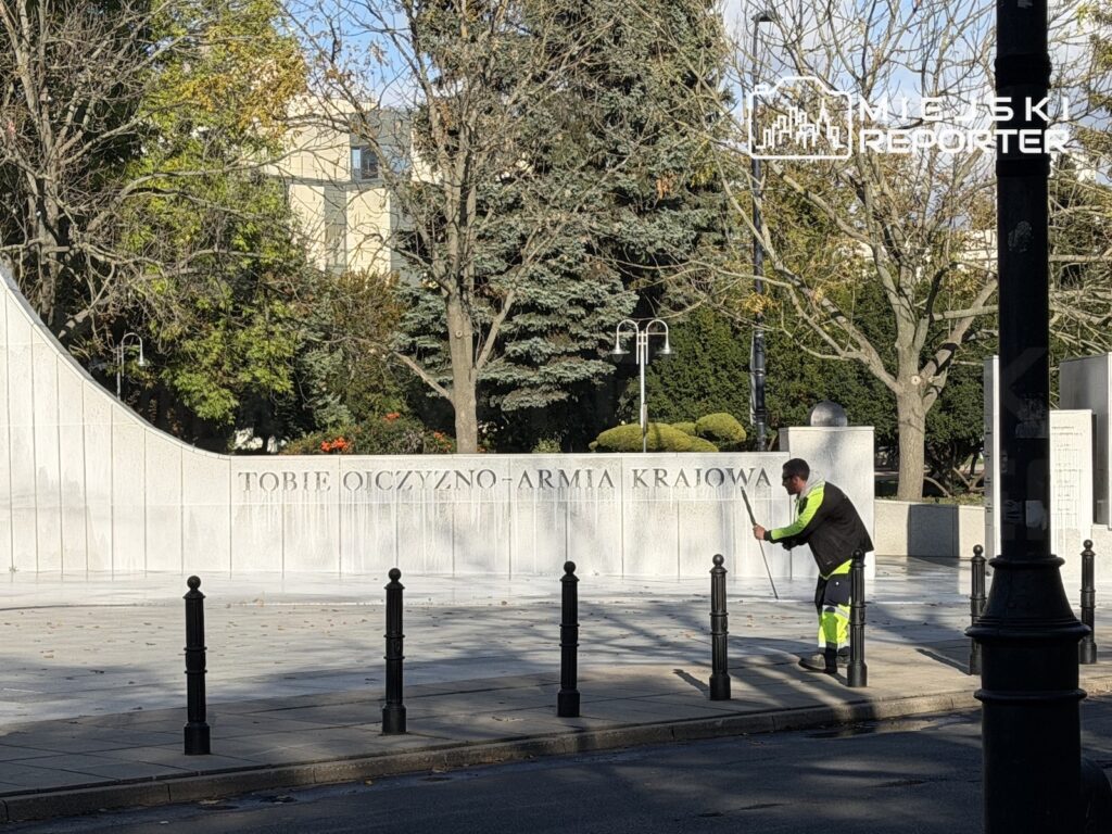 Pracownik w odblaskowym stroju czyści chodnik przed kamiennym pomnikiem z napisem „Tobie Ojczyzno - Armia Krajowa”.