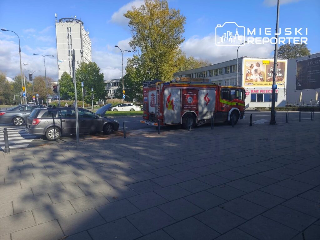 Czerwony wóz strażacki stoi przy ulicy obok zaparkowanych samochodów i chodnika.