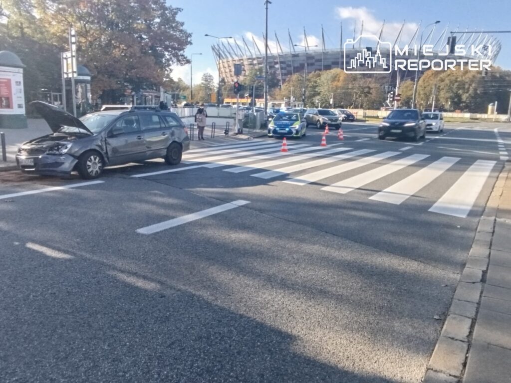 Uszkodzony samochód z otwartą maską stoi przy przejściu dla pieszych, a ruch zabezpieczają pachołki i radiowóz.