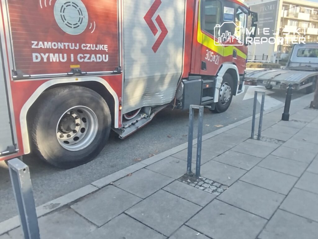 Czerwony wóz strażacki stoi przy chodniku; na boku widnieje napis „ZAMONTUJ CZUJKĘ DYMU I CZADU”.