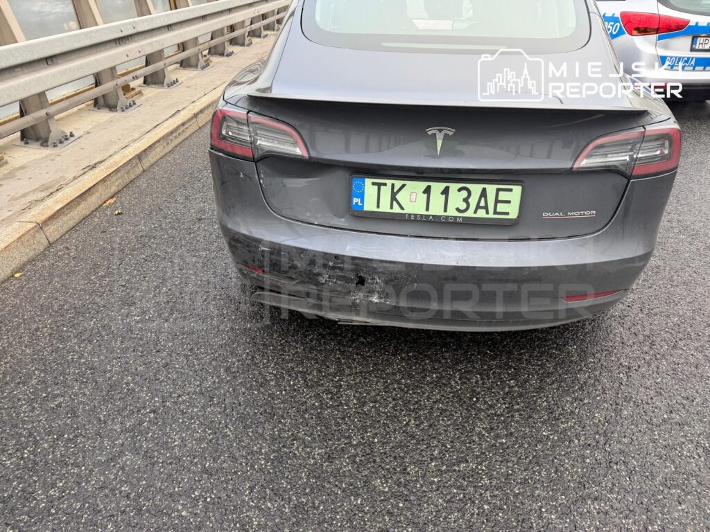 Tył szarej Tesli z uszkodzonym zderzakiem na drodze, obok barierki i policyjnego radiowozu.