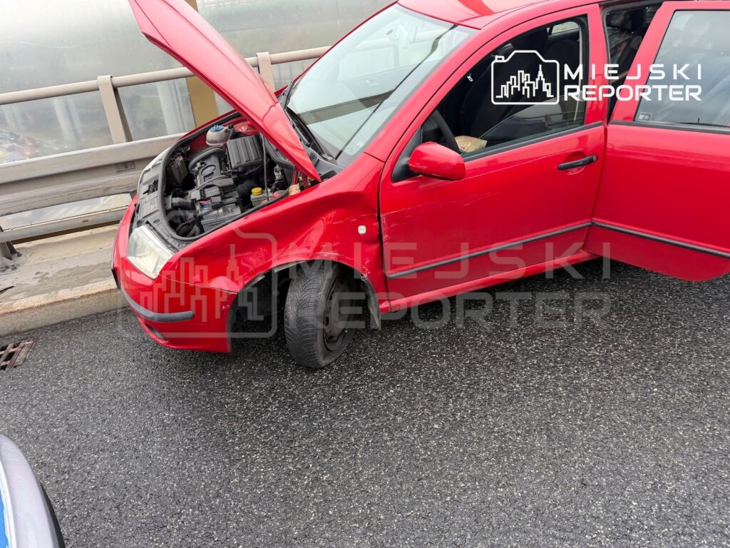 Czerwony samochód z otwartą maską i widocznym silnikiem stoi przy barierze drogowej.