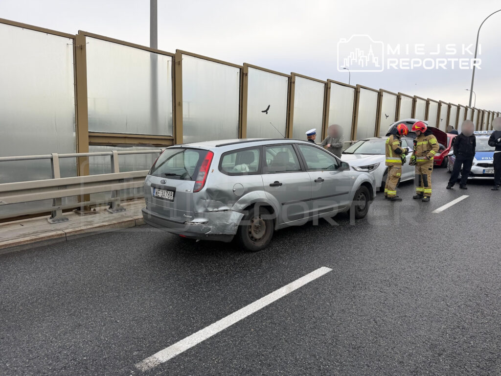 Uszkodzony srebrny samochód stoi na drodze przy strażakach i innych pojazdach po kolizji.