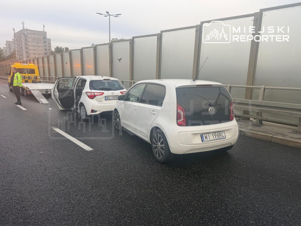 Dwa białe samochody stoją na pasie drogi, a obok nich żółta laweta i mężczyzna w kamizelce odblaskowej.