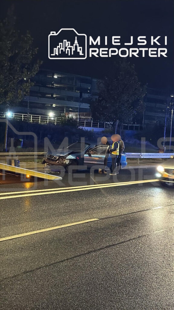 Uszkodzony przód samochodu na mokrej jezdni, obok stoją dwie osoby w kamizelkach odblaskowych.