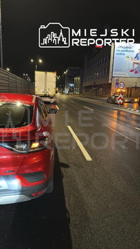 Czerwony samochód i biały ciężarowy jadą mokrą, oświetloną ulicą nocą, obok miejskich budynków.