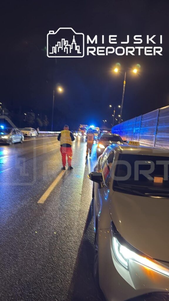 Uszkodzony samochód i służby na mokrej, nocnej jezdni; w tle migają niebieskie światła pojazdu ratunkowego.