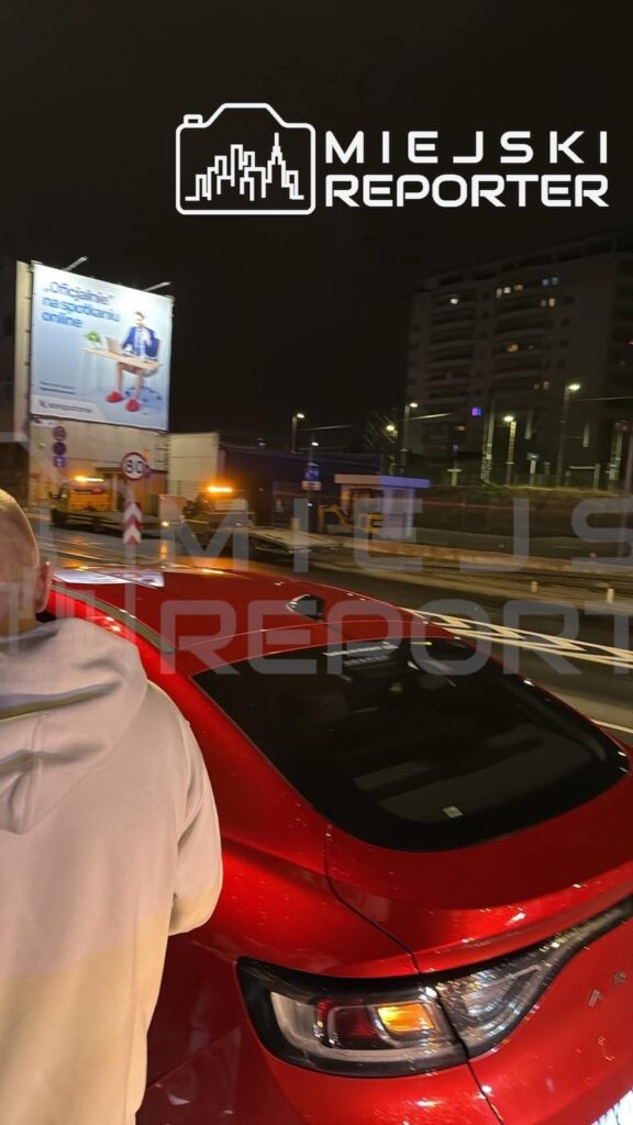 Czerwony samochód na nocnej ulicy, obok niego stojący człowiek i pojazd służb z włączonymi światłami ostrzegawczymi.