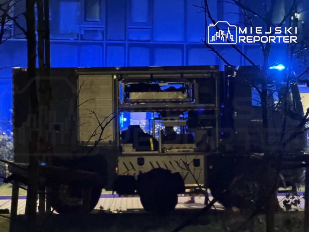 Ciężarówka z otwartą przestrzenią ładunkową stoi nocą przy niebiesko oświetlonym budynku; obok widać błysk niebieskiego