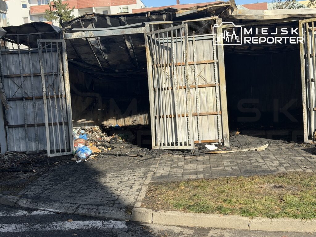 Nadpalona altana z otwartymi metalowymi bramami i śmieciami na podłodze, stojąca przy chodniku wśród zabudowy mieszkalnej.