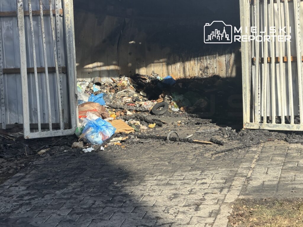 Zaśmiecone wnętrze otwartego, metalowego garażu z workami, odpadami i zabrudzoną posadzką.