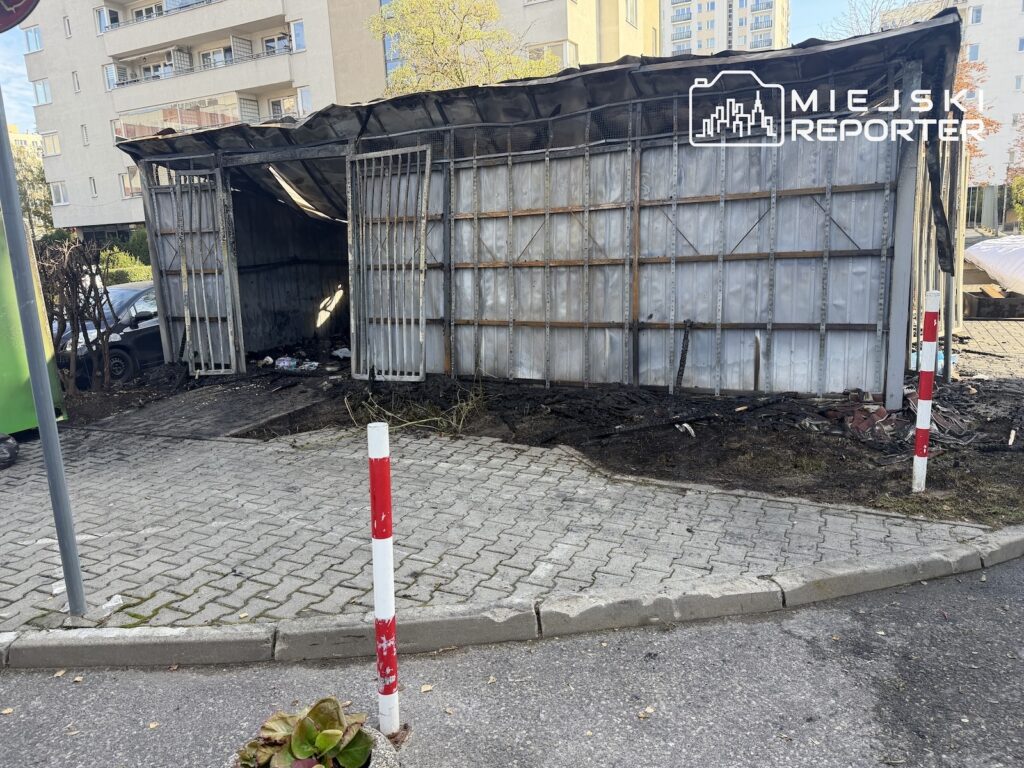 Osmalona, częściowo zniszczona drewniana wiata przy chodniku, z czarnymi śladami na ziemi i budynkami mieszkalnymi w tle.