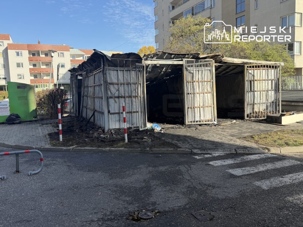 Spalone, uszkodzone wiaty śmietnikowe przy ulicy, na tle bloków mieszkalnych.