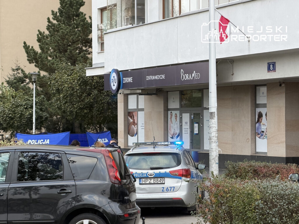 Radiowóz i samochód z oznaczeniami policji stoją przed budynkiem z centrum medycznym, za policyjnymi parawanami.