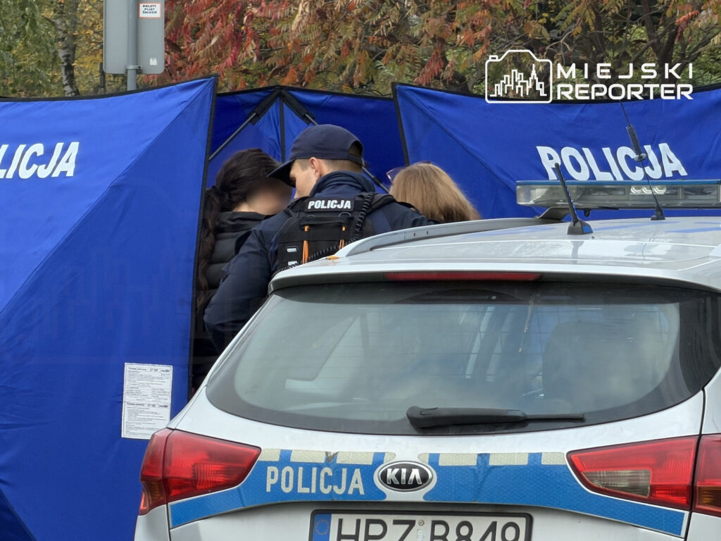 Policjanci za niebieskimi parawanami z napisem „POLICJA” obok radiowozu, na tle jesiennych drzew.
