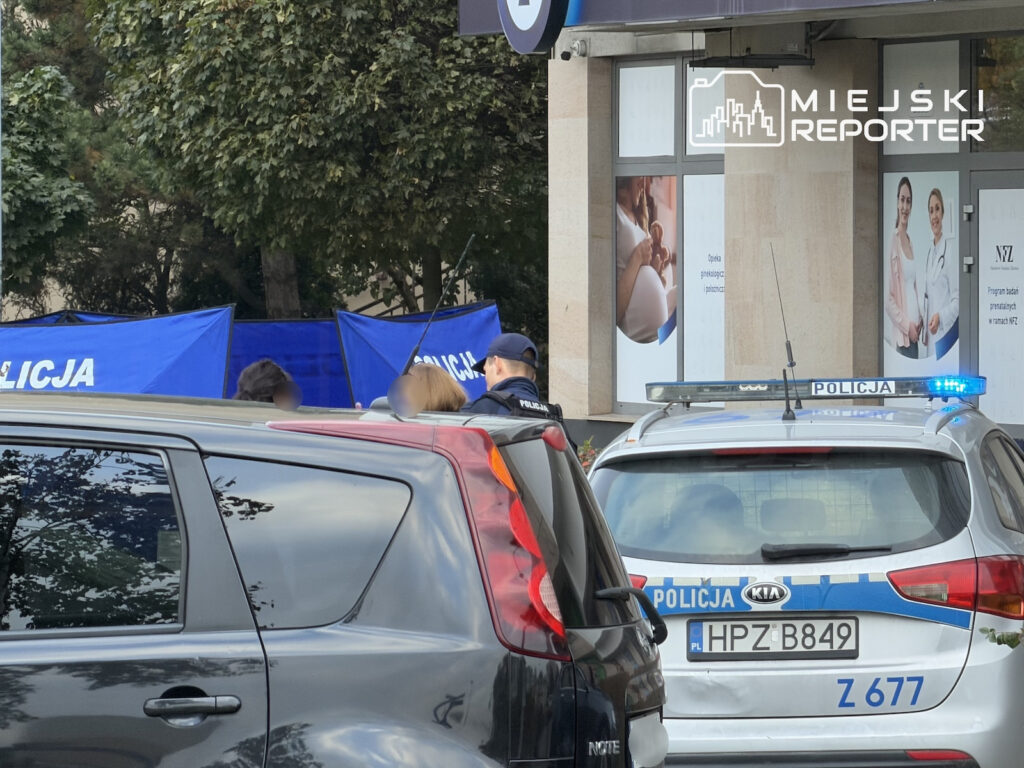 Radiowóz i funkcjonariusze policji przy niebieskich parawanach przed budynkiem mieszkalnym w Ursynowie.