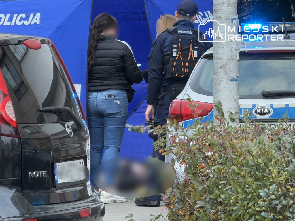 Policjant i kobieta stoją przy niebieskim parawanie policyjnym między zaparkowanymi samochodami; na chodniku leży rozmyta